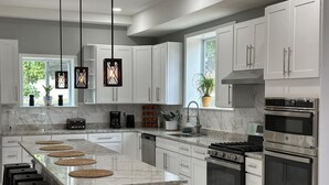 A modern kitchen with white cabinets, a large island with stools, stainless steel appliances, and pendant lighting. - Caton Manor Estate and Pool (Catonsville)