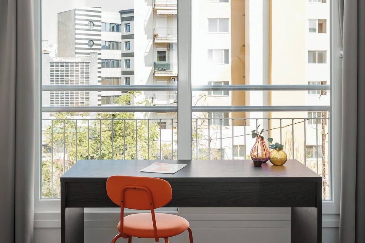 A modern room with a dark wooden desk, a red chair, and a view of a cityscape through large windows.