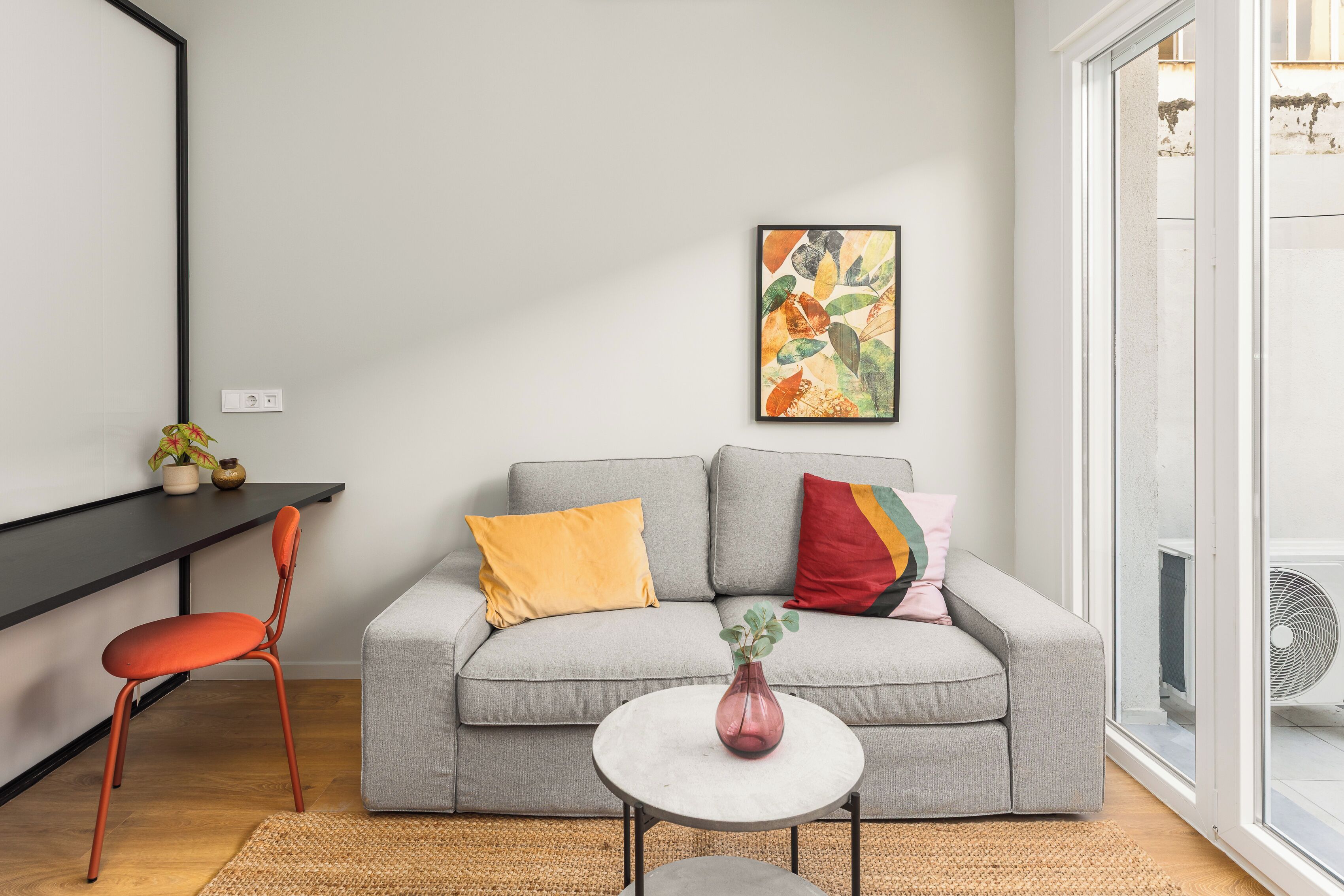 Una sala de estar moderna con un sofá gris, un sillón rojo, una mesita redonda y un cuadro abstracto de colores en la pared.