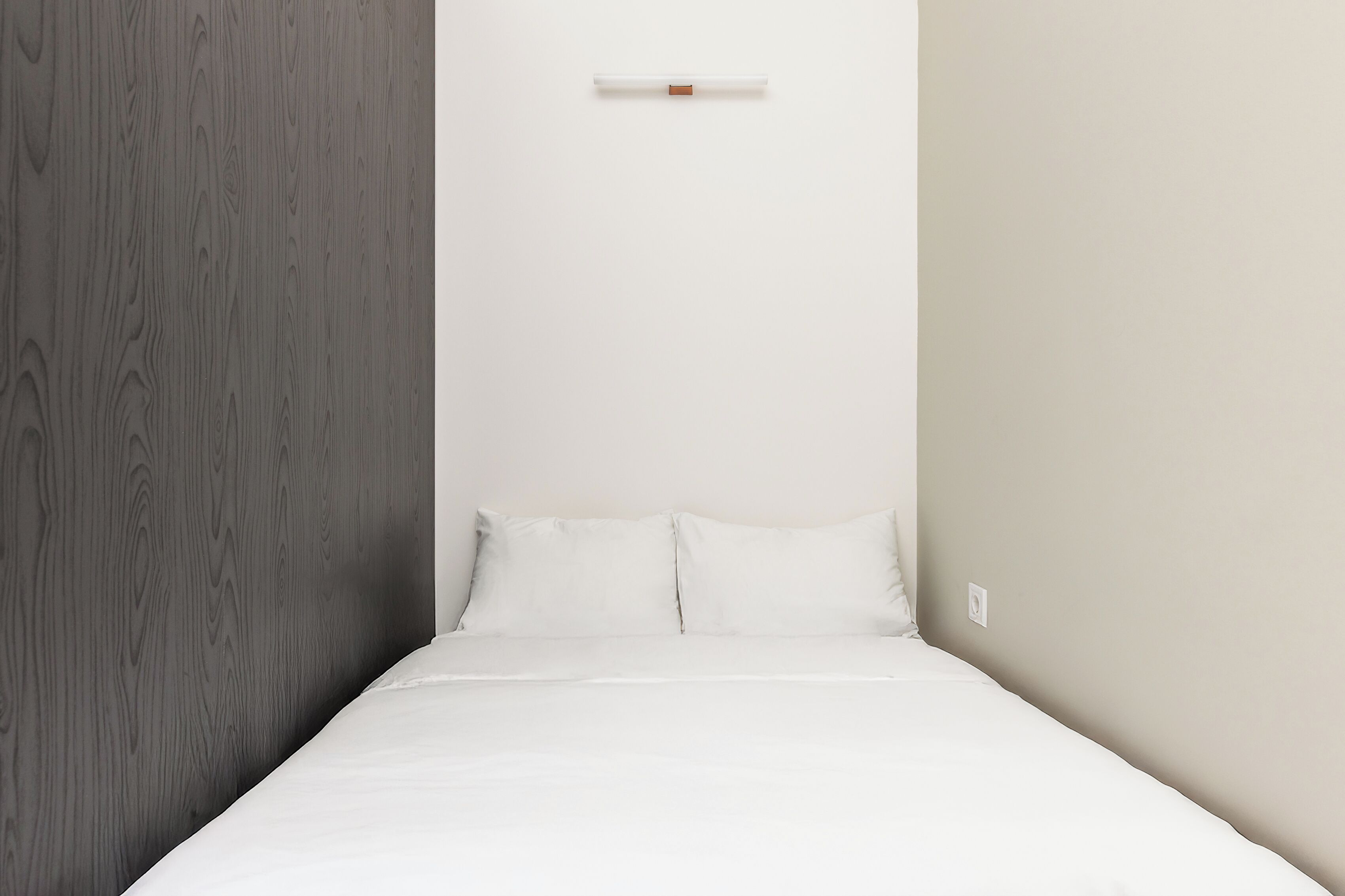 A minimalist hotel room with a bed, white bedding, a wooden accent wall, and a plain white wall.