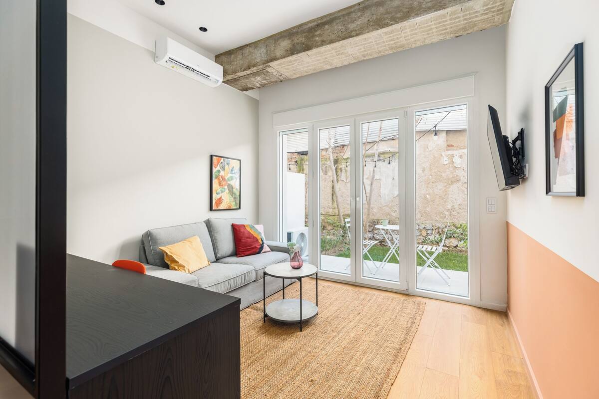 A modern living room with a sofa, a coffee table, and a TV. There is a large window with a view of the outside.