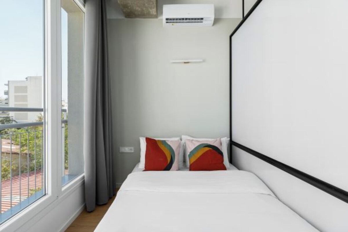 Una habitación de hotel moderna con un ventanal grande, una cama con ropa de cama blanca y almohadas coloridas.