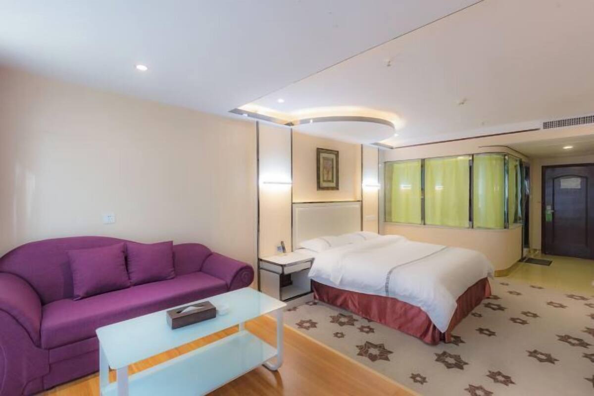Una habitación de hotel con una cama, un sofá morado, una mesita, una mesita de noche y una ventana con cortinas.