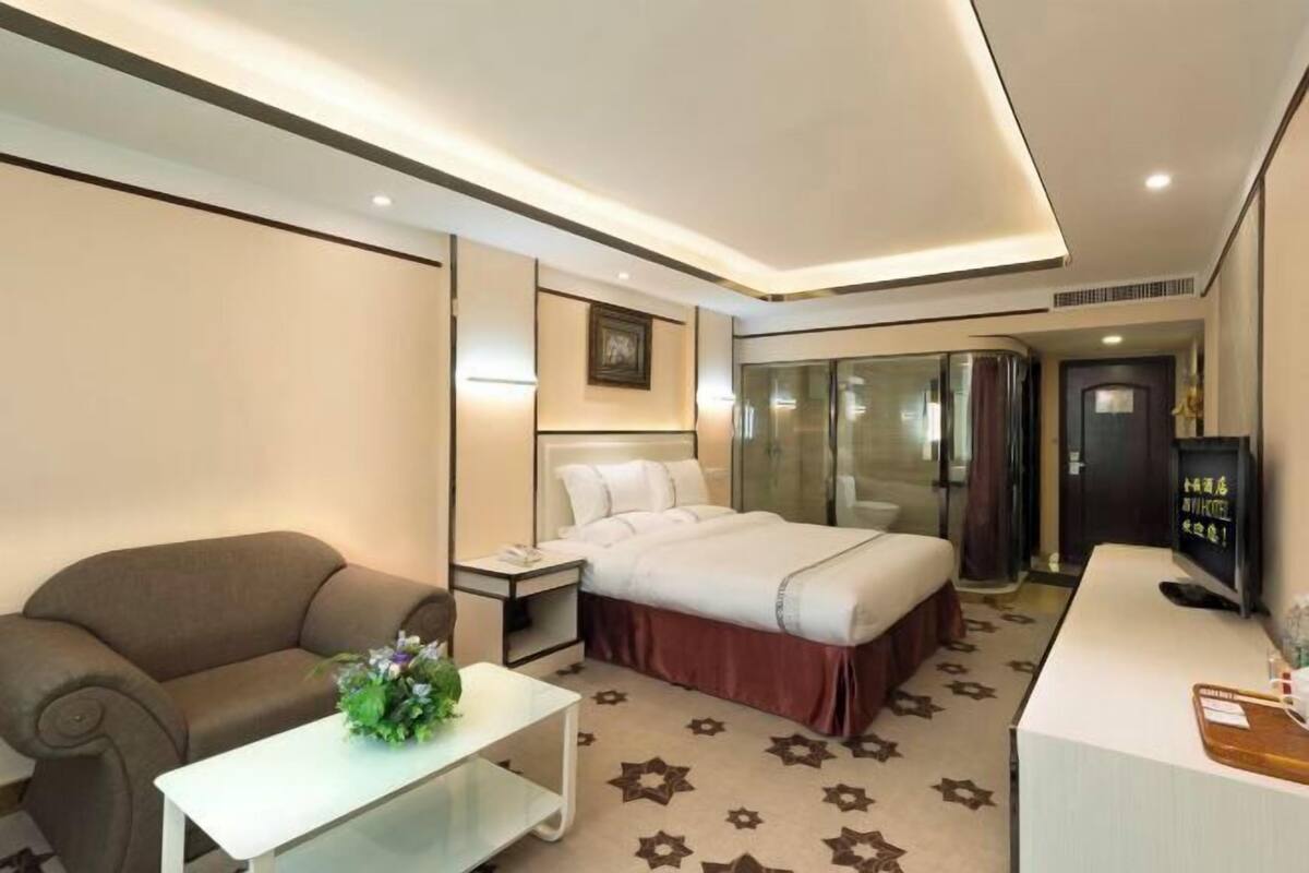 Una habitación de hotel con cama, sofá, una mesita con un florero, televisor y escritorio con ordenador.