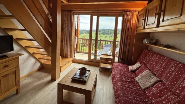 Een houten blokhut van binnen met een bank, een tafel, een televisie en een balkon met uitzicht op het landschap.