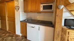 Een compacte keuken met houten kastjes, een magnetron, een vaatwasser en een kleine televisie op een houten standaard.
