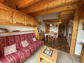 A cozy cabin interior with a red patterned sofa, wooden dining table, and a kitchen area with wooden cabinets and a microwave.