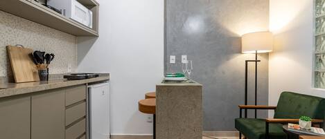 A compact kitchen with a microwave, a small refrigerator, and a countertop with a cutting board and utensils. There is a green chair and a table with a lamp and a small plant.