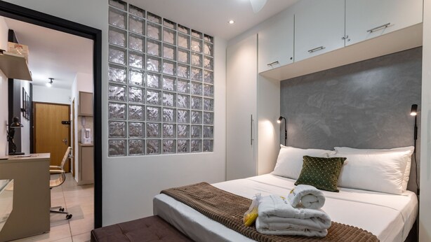A modern hotel room with a large bed, a bench, a glass block wall, and a desk with a chair. - In Front of the Underground and 8 Minutes From the Beach Sf228 (Rio de Janeiro)
