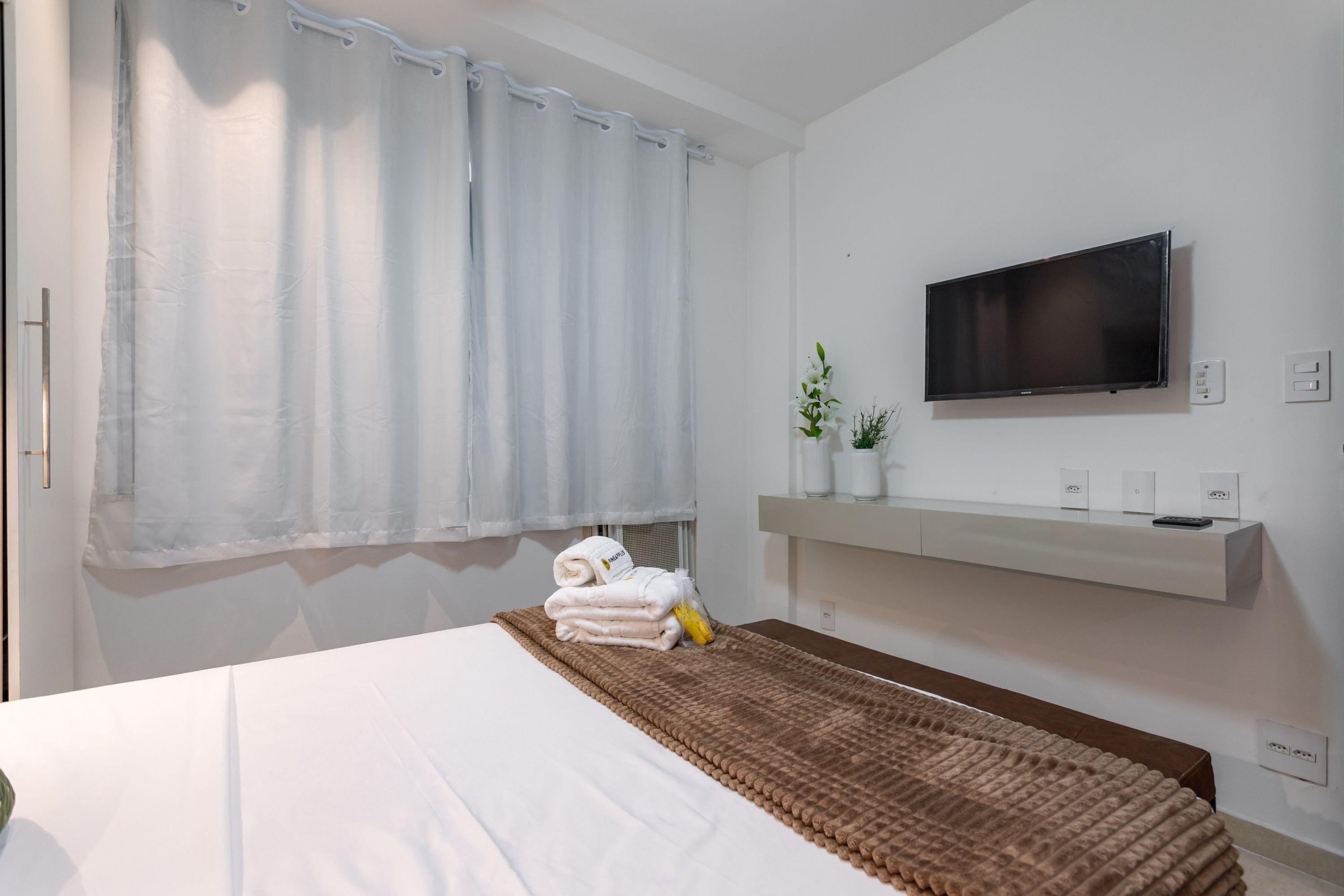 Une chambre d’hôtel avec un lit, un téléviseur fixé au mur, une fenêtre avec un rideau et une petite plante posée sur une étagère.