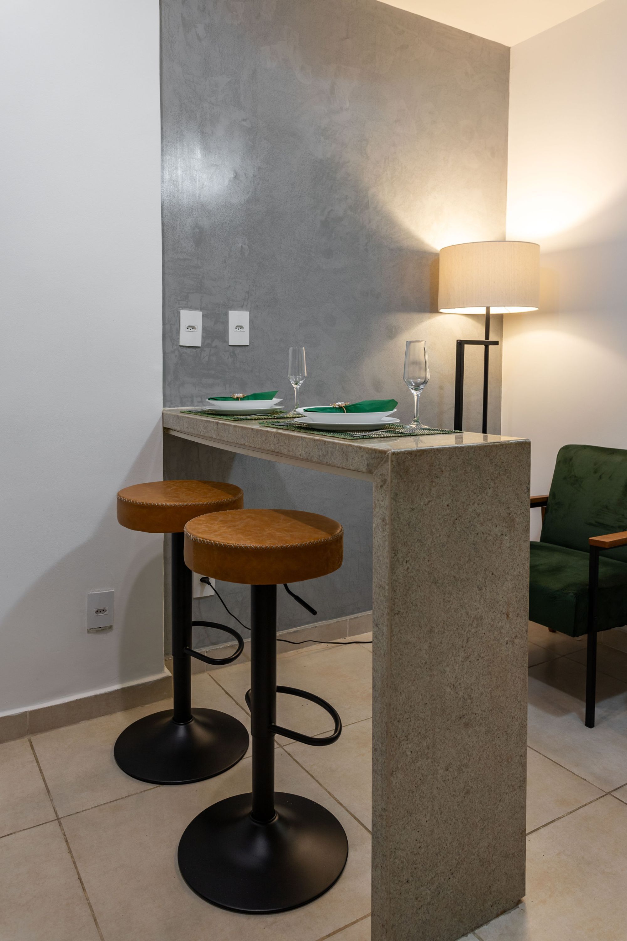 A modern kitchen with a granite countertop, two stools, a green chair, and a table lamp.