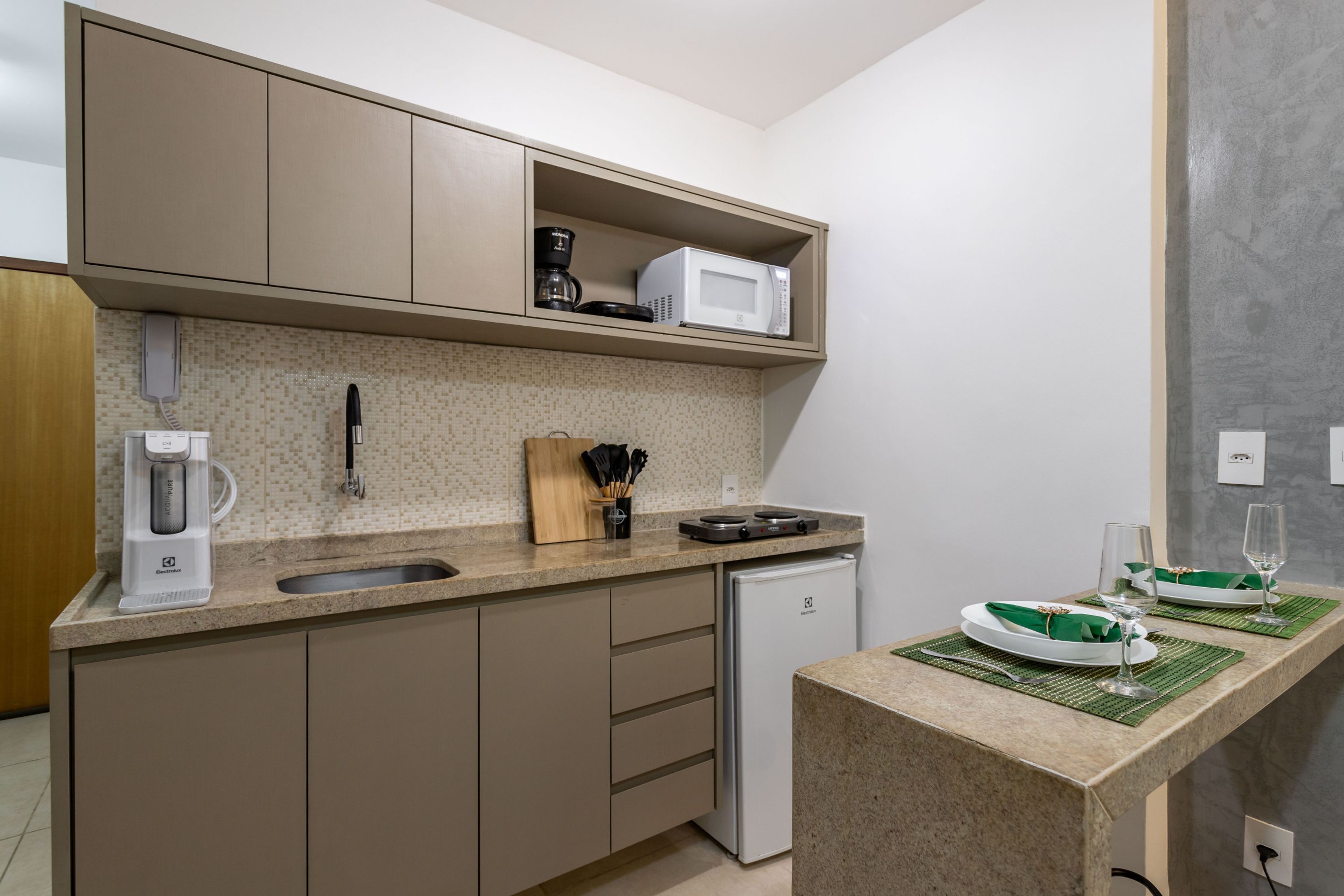 Cocina compacta con cafetera, microondas y un refrigerador pequeño. Hay una encimera con un lavamanos y una zona de comedor con una mesa para dos.