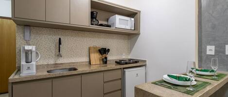 Cocina compacta con cafetera, microondas y un refrigerador pequeño. Hay una encimera con un lavamanos y una zona de comedor con una mesa para dos.