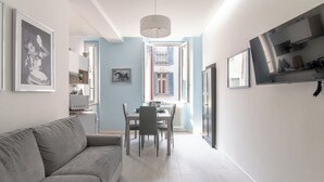 Una sala moderna con un sofá gris, una mesa de comedor con sillas y una cocina visible a través de una puerta abierta.