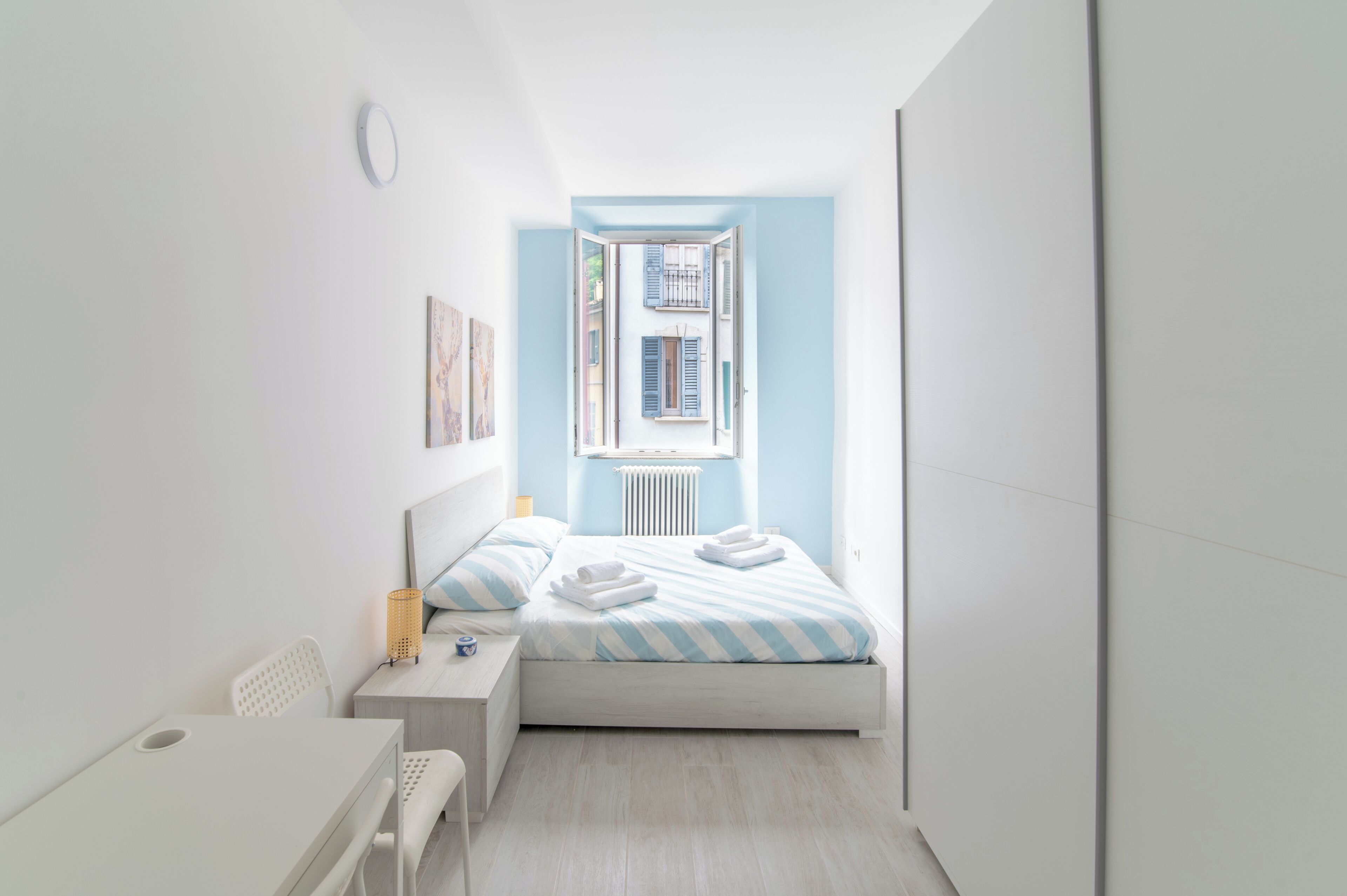 A small, bright room with a bed, a bedside table, a chair, and a window with shutters.