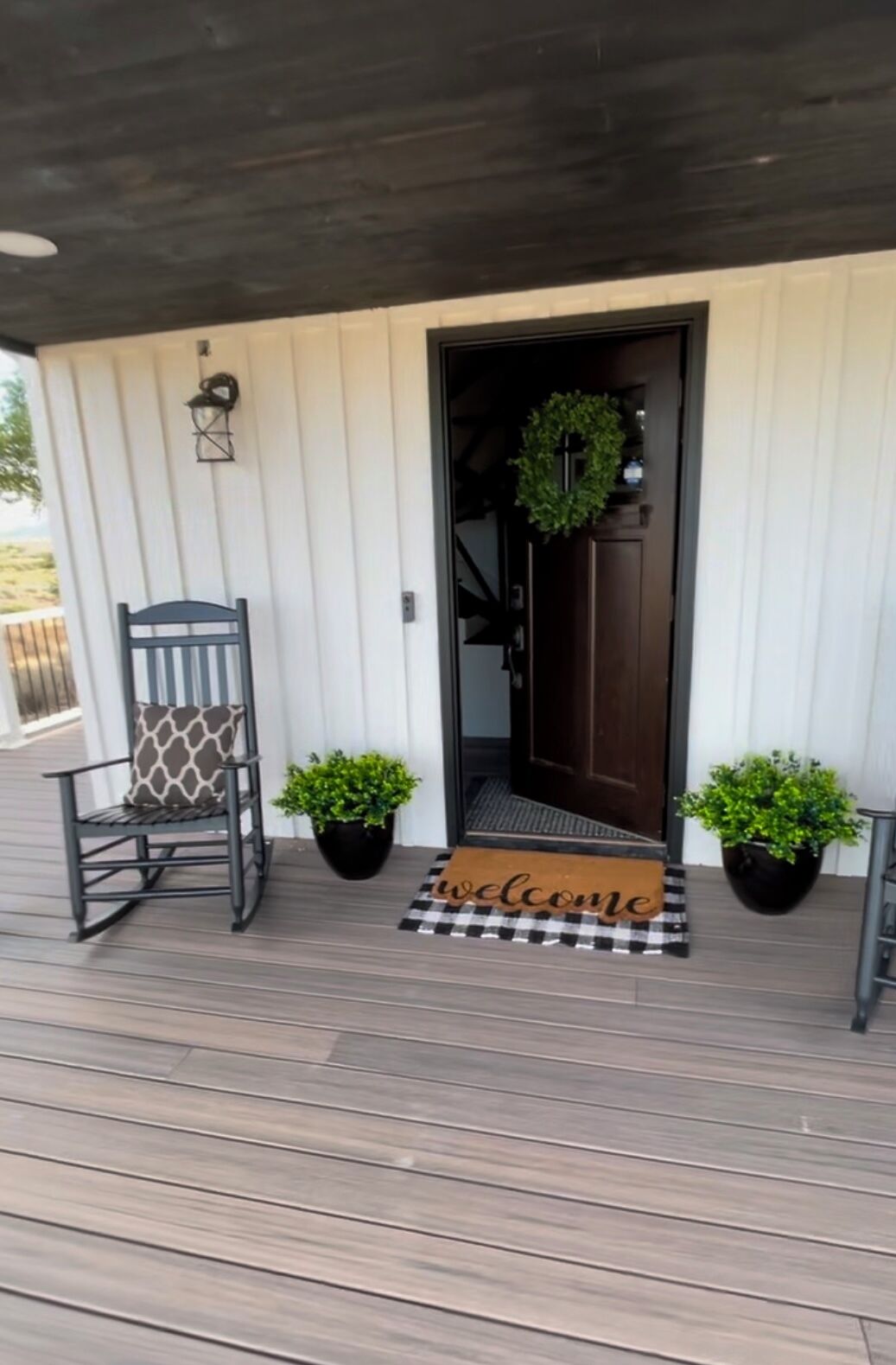 A wooden deck with a dark brown door, a welcome mat, and two potted plants.