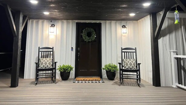 A covered porch with two wooden chairs and potted plants, a dark door with a wreath, and a welcome mat. - Escape to Luxurious Lynndyl Resort—Nature, Comfort & Serenity Await (Lynndyl)