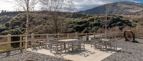 Un área de estar al aire libre con mesas y sillas, rodeada por una valla de madera, con colinas y un cielo nublado de fondo.