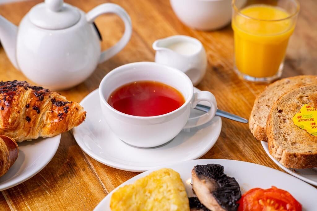 A breakfast setting with croissants, a cup of tea, toast, a glass of orange juice, and a teapot on a wooden table.