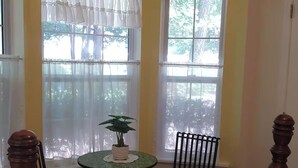 A room with a bed, two chairs, a small table with a plant, and two windows with sheer curtains.