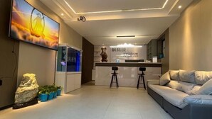A modern living room with a sofa, a TV mounted on the wall, a kitchen area with a counter and stools, and a decorative rock and plants on the floor. - Altay Fenglinwan Boutique Hotel (Altay City)