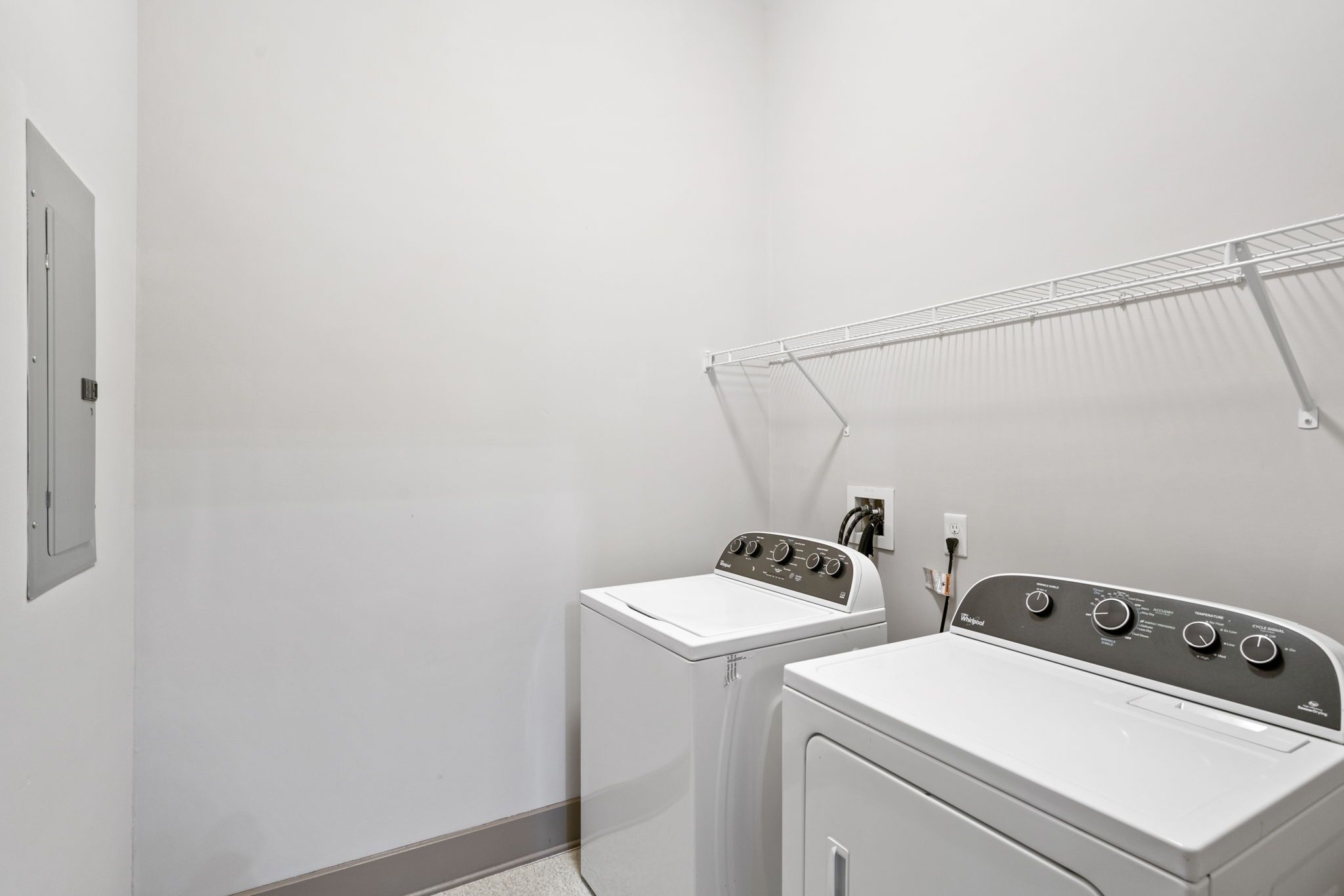 A laundry room with two white washers and dryers, a metal drying rack, and a wall-mounted electrical outlet.