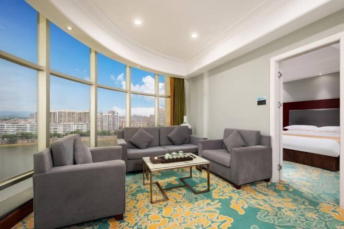 Ein Hotelzimmer mit großem Fenster und Blick auf die Stadt, einem Sofa, einem Sessel, einem kleinen Tisch und einem Bett.