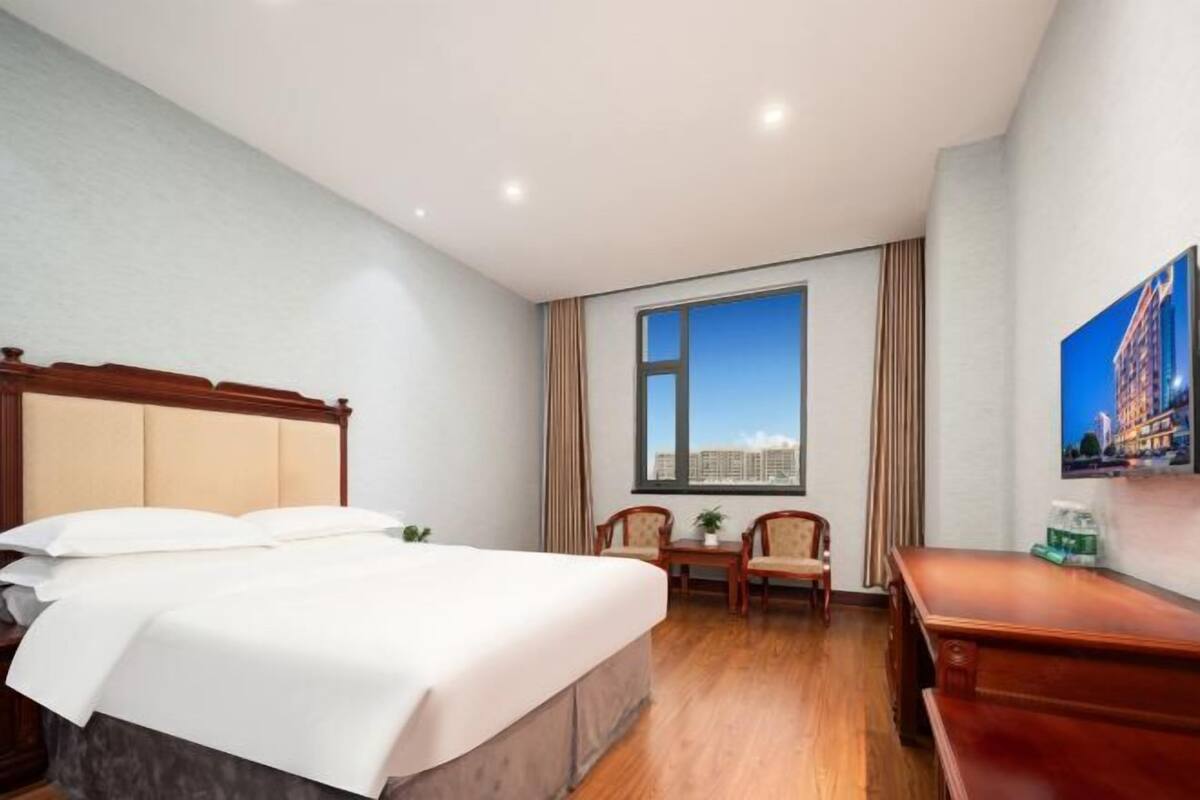 Ein Hotelzimmer mit einem großen Bett, einem Schreibtisch, zwei Stühlen, einem Fenster mit Stadtblick und einem an der Wand befestigten Fernseher.