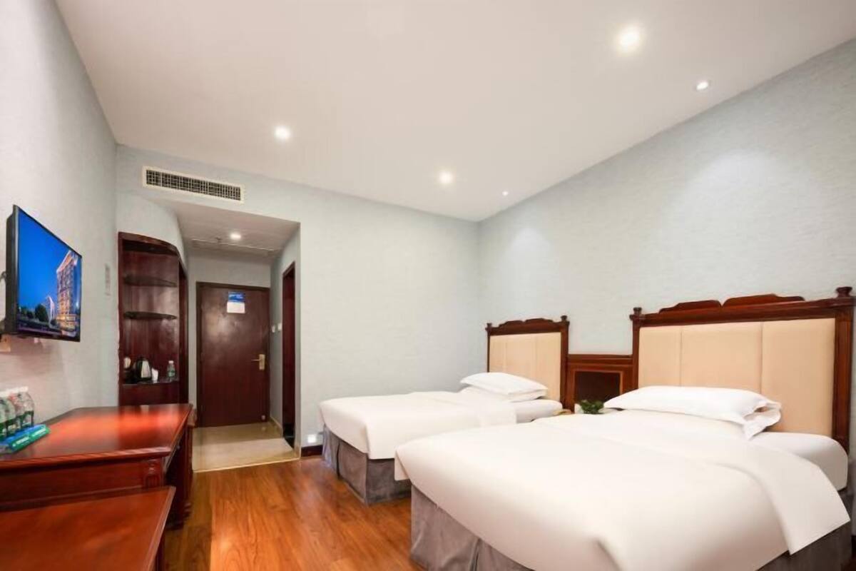Ein Hotelzimmer mit zwei Einzelbetten, einem Holztisch, einem Fernseher und einer Tür zu einem weiteren Raum.