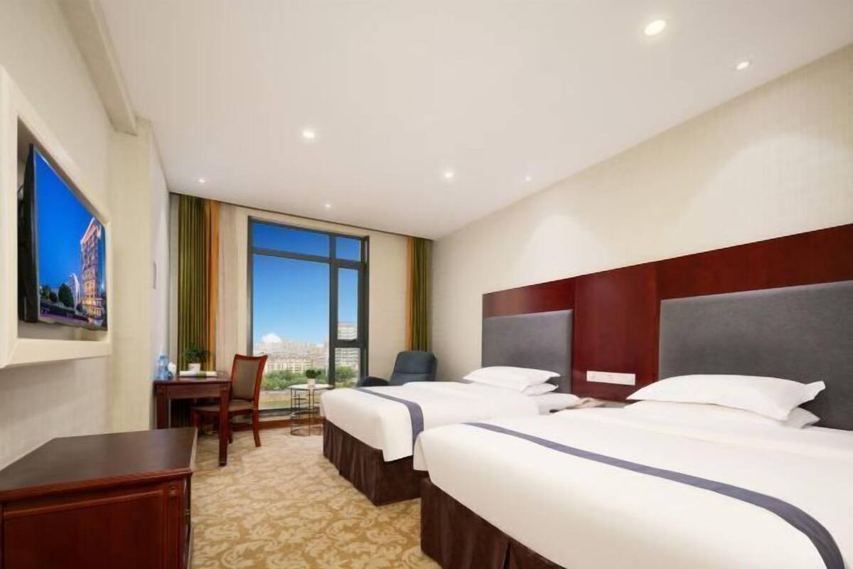 Ein Hotelzimmer mit zwei Betten, einem Schreibtisch, einem Sessel und einem großen Fenster mit Blick auf die Stadt.