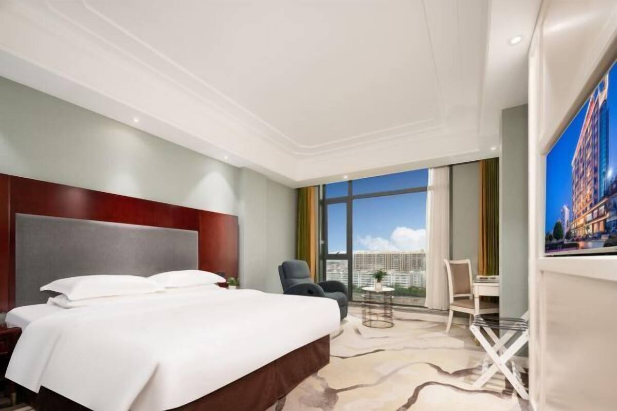 Ein Hotelzimmer mit einem großen Bett, Blick auf die Stadt durch ein großes Fenster, einem kleinen Tisch und einem Sessel.
