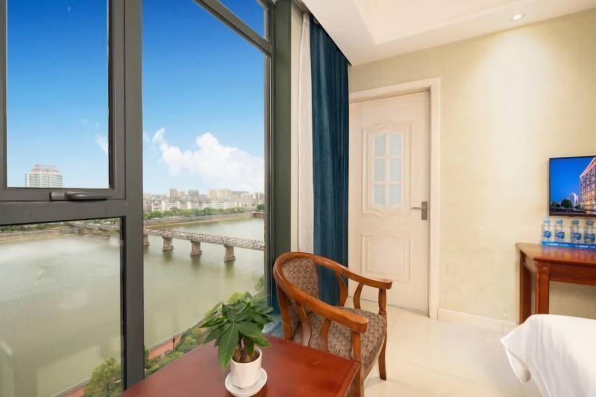 Ein Hotelzimmer mit großem Fenster und Blick auf einen Fluss und eine Brücke, ein Holzstuhl, ein kleiner Tisch mit Topfpflanze sowie ein Fernseher auf einem Holzgestell.
