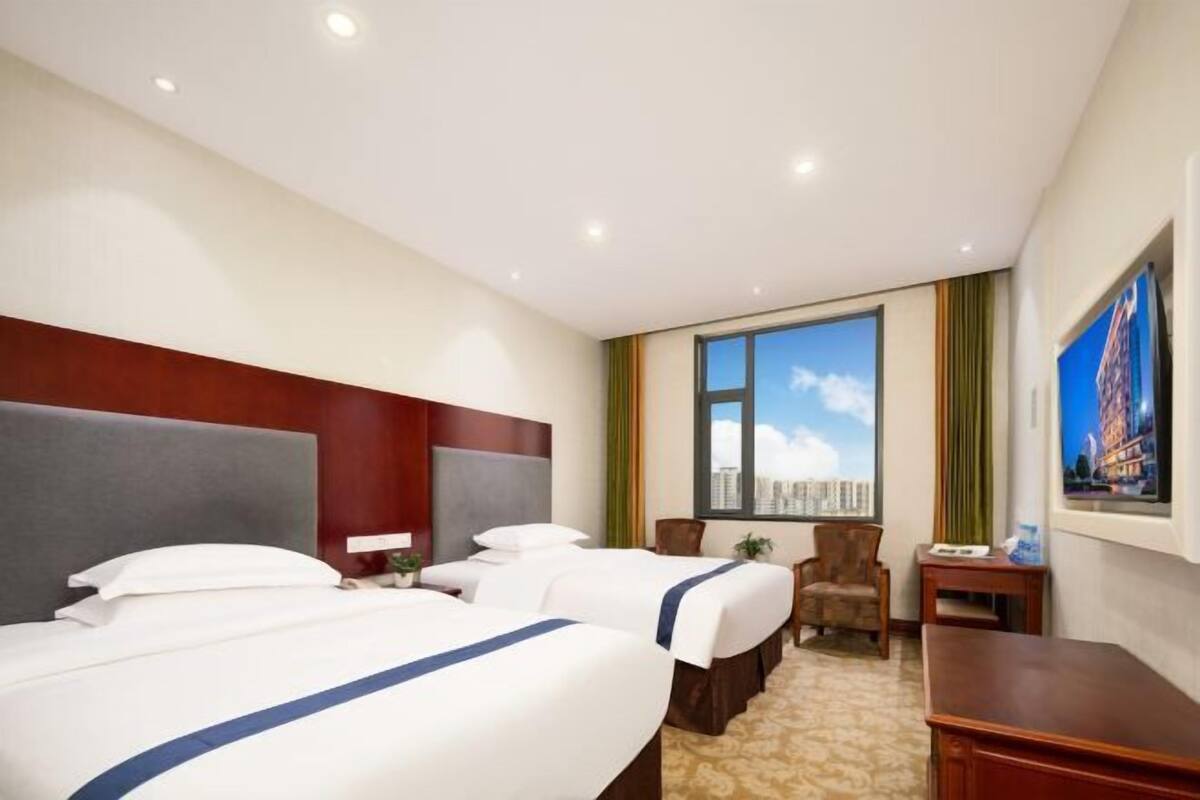 Ein Hotelzimmer mit zwei Betten, einem Schreibtisch, einem Stuhl, einem Fenster mit Blick auf die Stadt und einem Bild an der Wand.