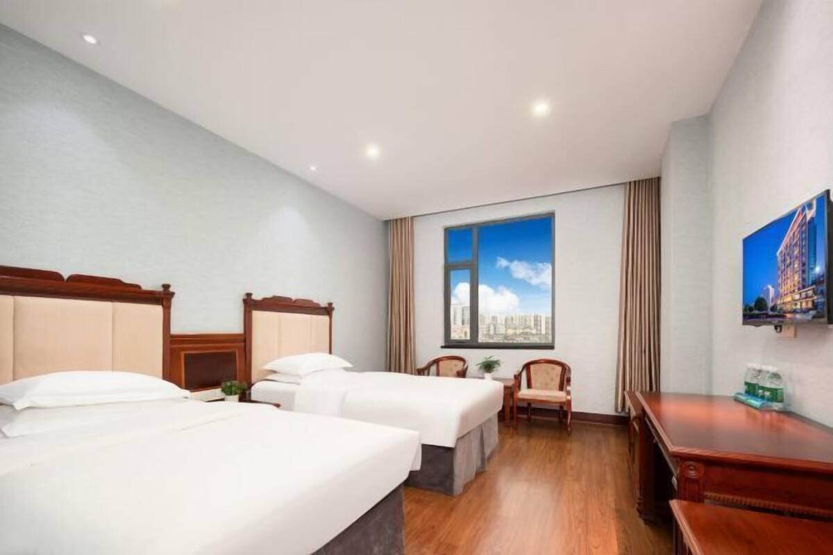 Ein Hotelzimmer mit zwei Betten, einem Schreibtisch, einem Stuhl und einem großen Fenster mit Blick auf die Stadt.