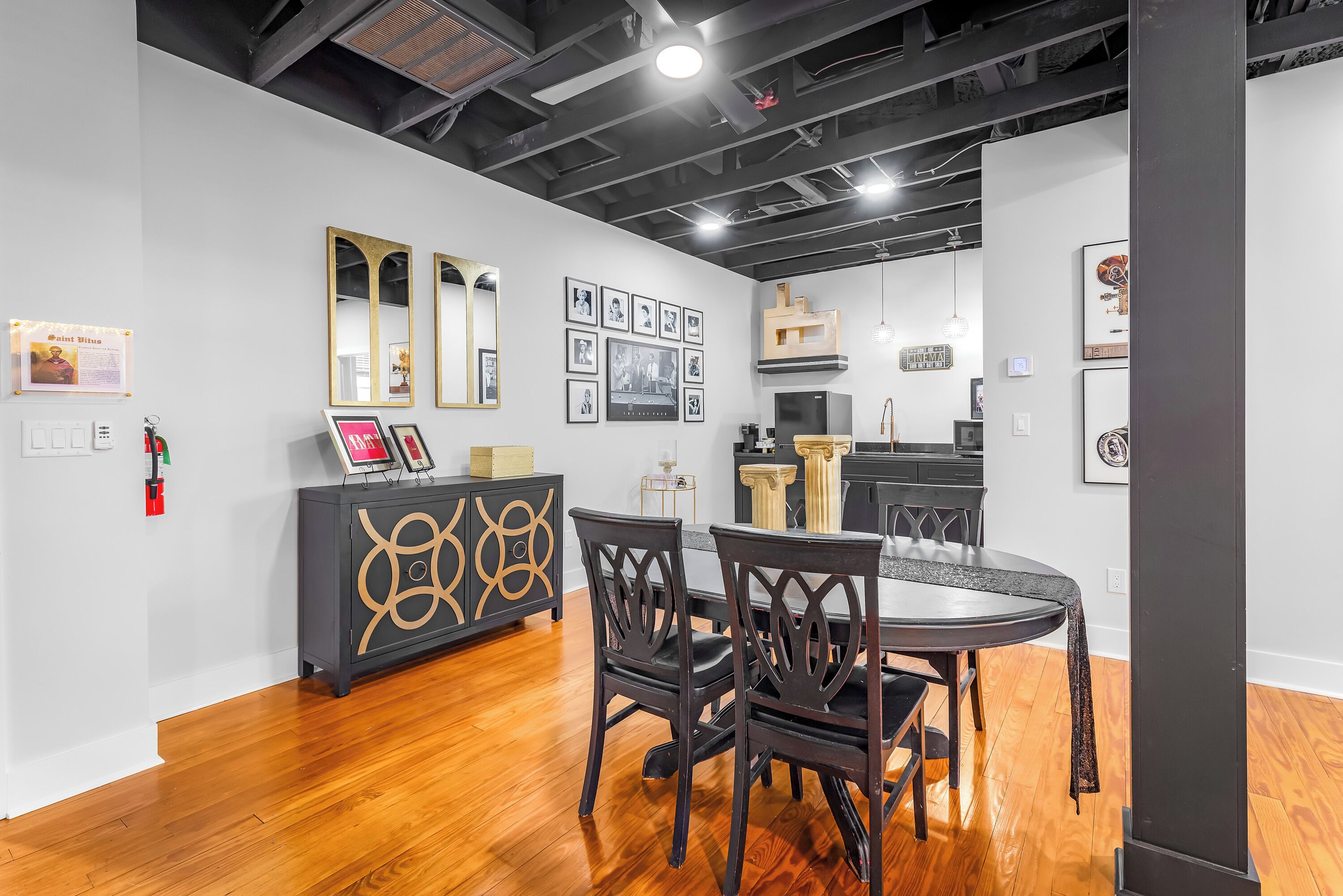 Un área de comedor moderna con suelo de madera, sillas de madera oscura y una mesa redonda. Hay cuadros en la pared y un mueble negro con diseños decorativos.