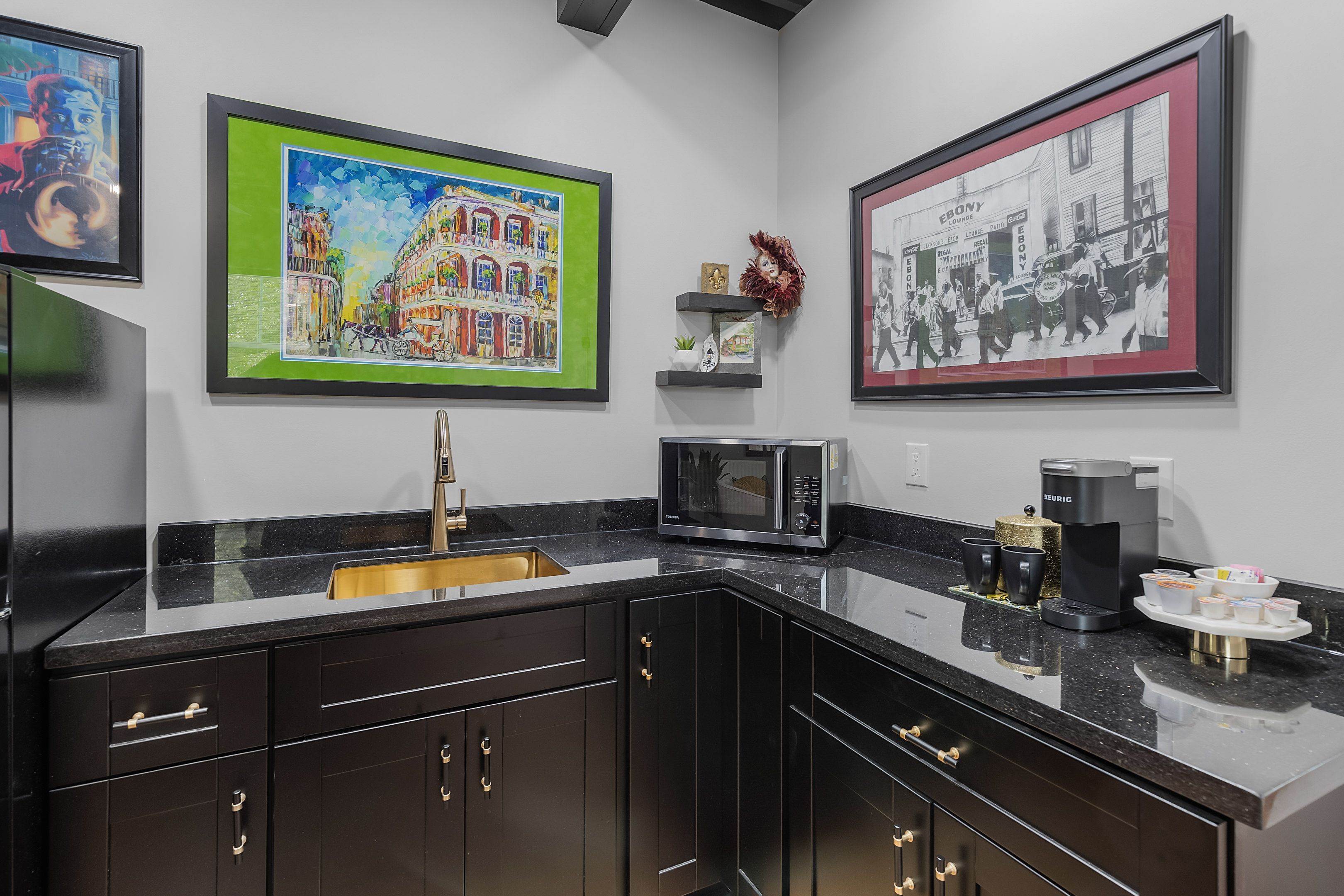 Een moderne keuken met donkere kastjes, een zwart aanrecht, een roestvrijstalen spoelbak, een magnetron en een koffiezetapparaat. Er hangen kunstwerken in lijsten aan de muur boven het aanrecht.