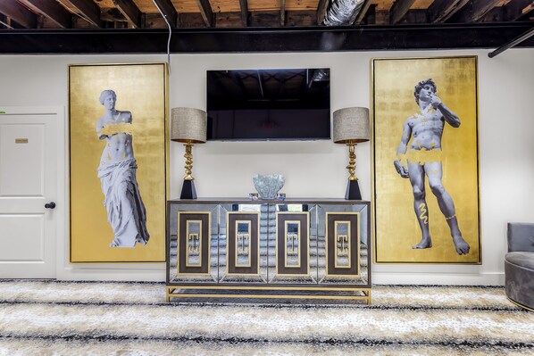 A room with a large gold-framed artwork on the wall, a flat-screen TV above a console table, and a patterned carpet.