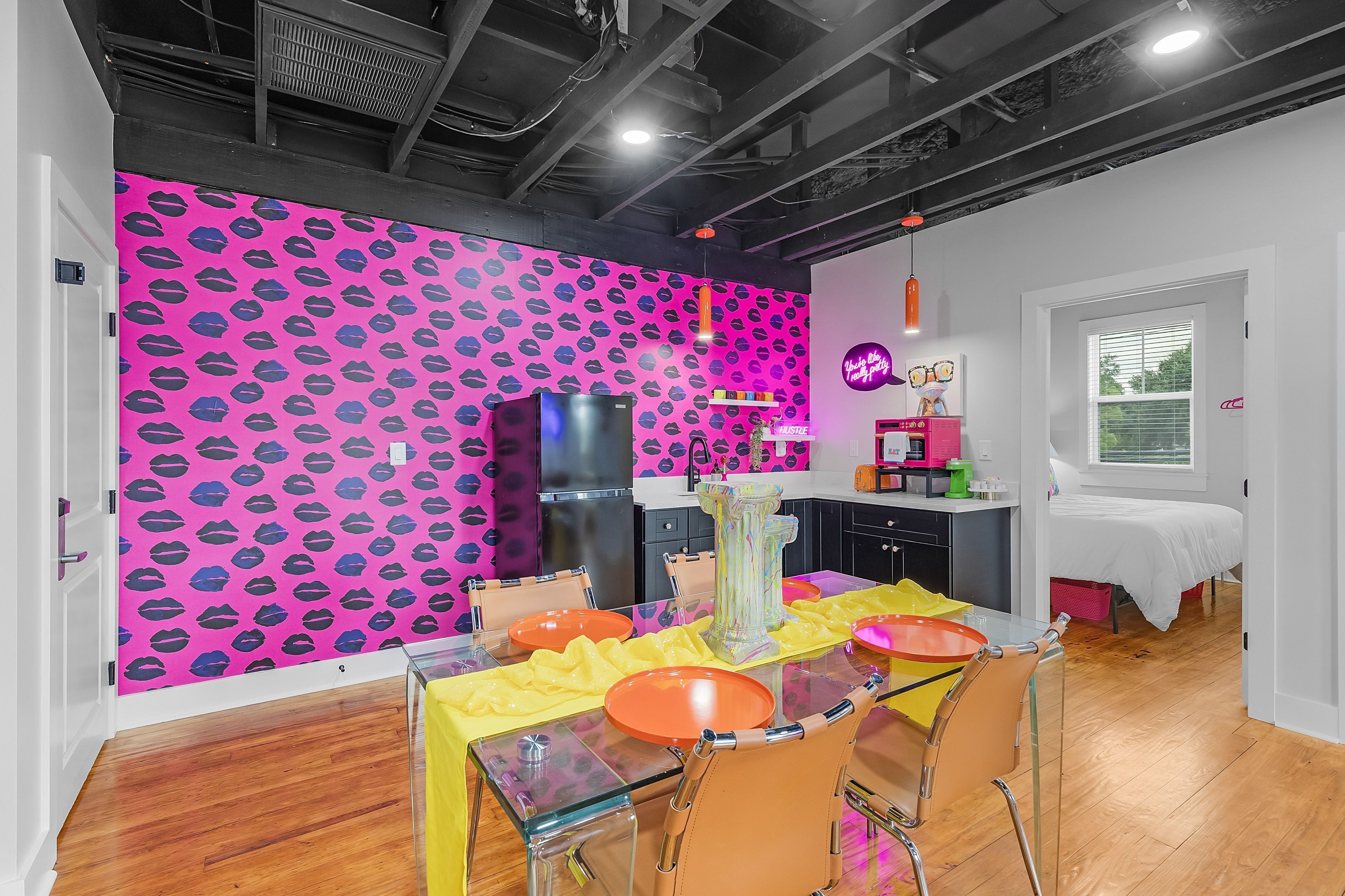 Una cucina moderna con una parete rosa, un'area pranzo con tavolo in vetro e un letto sullo sfondo.