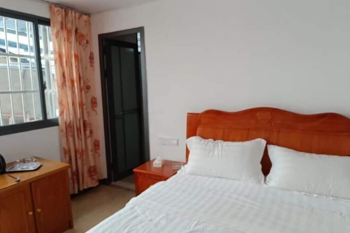 Una habitación de hotel con cama, mesita de noche de madera y ventana con cortinas.