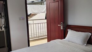 Una habitació d'hotel amb un llit, un panell de capçalera de fusta, una porta i vista a un balcó amb una cortina blanca.