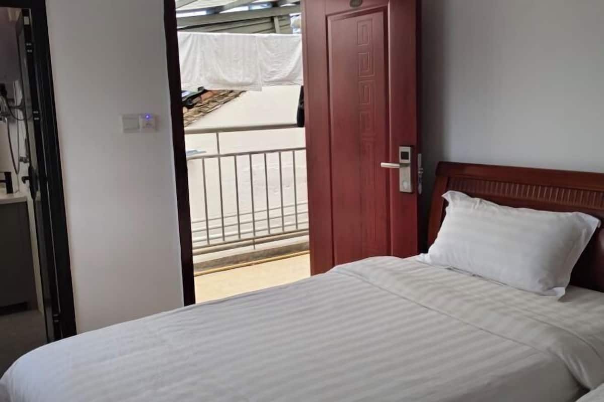 Habitación de hotel con cama, cabecera de madera, una puerta y vistas a un balcón con cortina blanca.