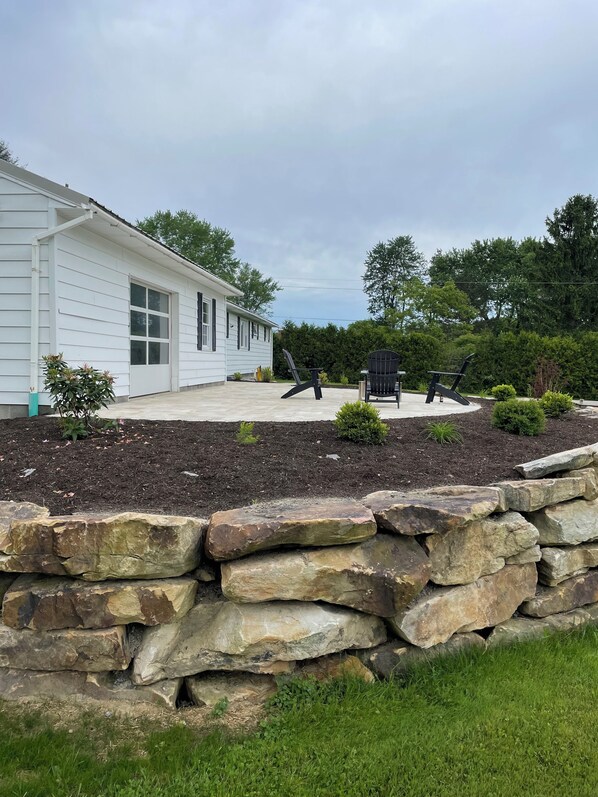 A white house with a stone retaining wall, a patio with outdoor furniture, and a garden with various plants. - 3-bedroom house with spacious private yard and fire pit in Saint Marys (Saint Marys)