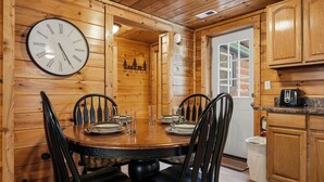 Un interno in legno con un tavolo rotondo, sedie nere, una sveglia a parete e un angolo cucina con mobili in legno.
