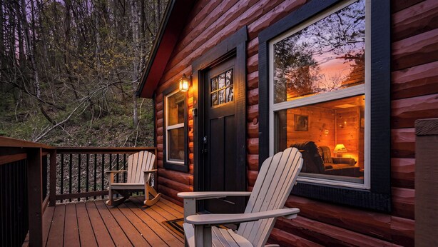 Una baita di tronchi con una veranda, due poltrone Adirondack e un interno illuminato visibile attraverso le finestre.