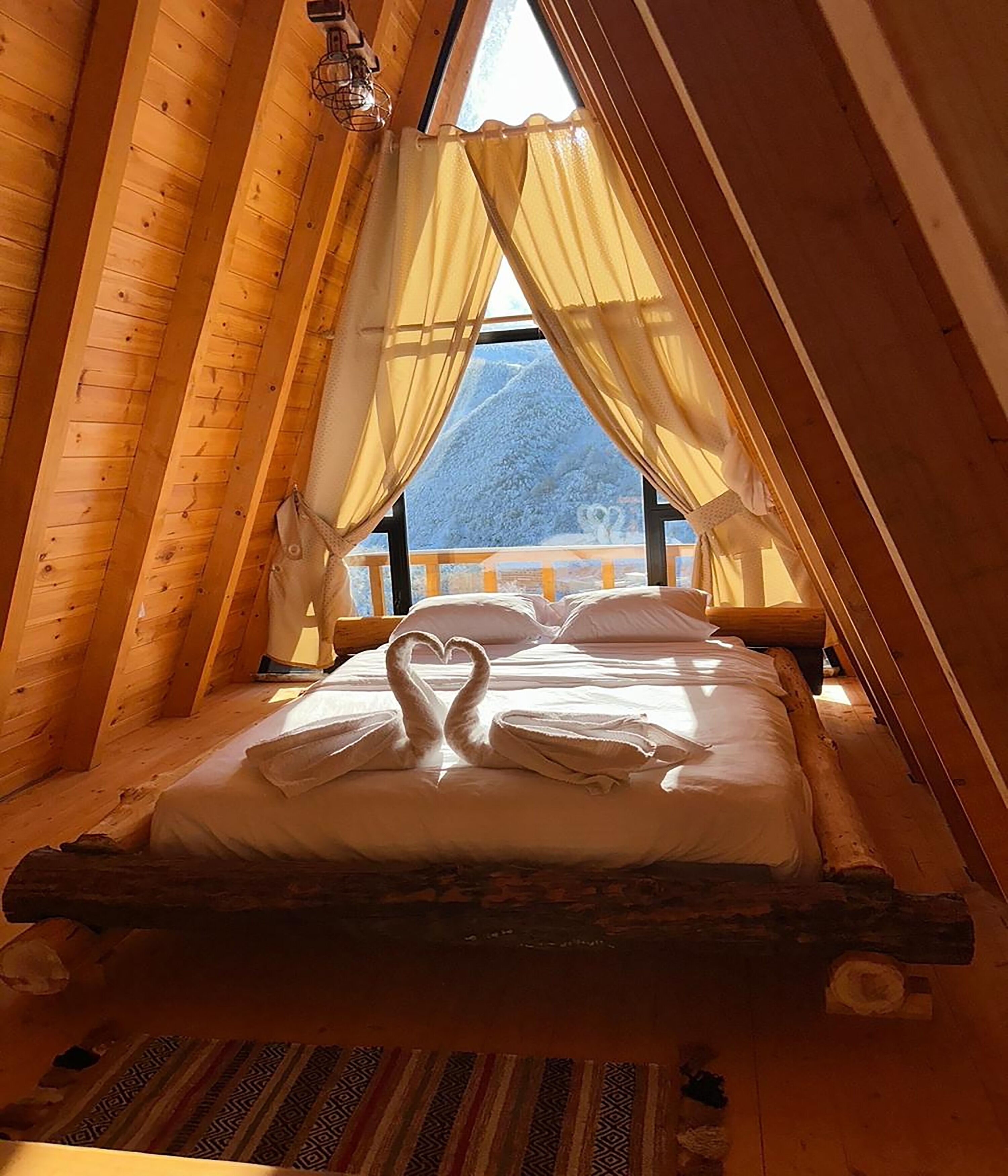 A cozy wooden cabin bedroom with a bed, a heart-shaped pillow arrangement, and a view of a snowy mountain.