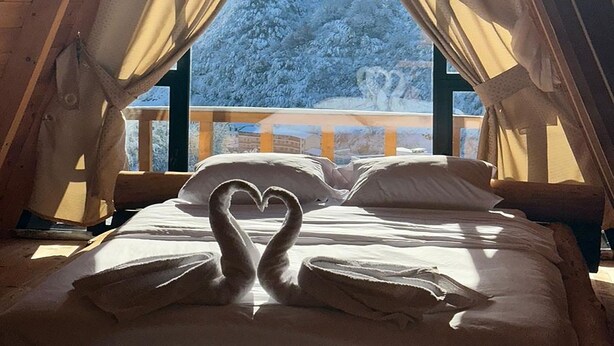Una cama acogedora con una disposición de almohadas en forma de corazón, vigas de madera y vistas a un paisaje nevado a través de cortinas translúcidas.