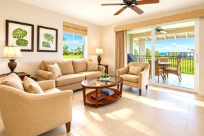 Habitación de hotel con sofá, sillones, una mesa de centro, balcón con vista al océano y dos cuadros con plantas tropicales en la pared.