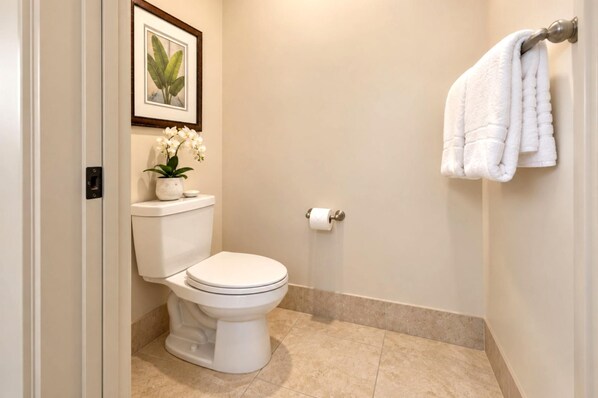 A bathroom with a toilet, a framed plant, a towel rack, and a toilet paper holder.