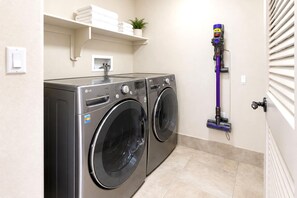 Une buanderie équipée de deux lave-linge LG à chargement frontal, d’un aspirateur fixé au mur et d’une étagère où sont rangées des serviettes pliées et une plante en pot.