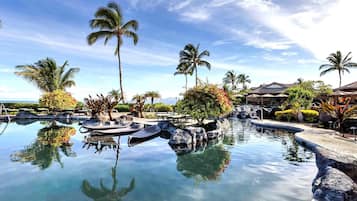 A pool with clear water, surrounded by rocks and palm trees, with lounge chairs and umbrellas in the background.